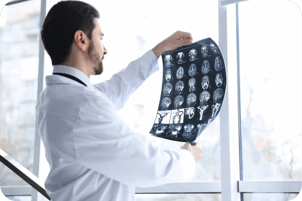 A imagem mostra um médico, de jaleco branco, em um ambiente bem iluminado (provavelmente um consultório ou clínica, com uma grande janela ao fundo), olhando atentamente para uma folha de exame de imagem. A folha, que parece ser uma chapa de ressonância magnética ou tomografia, exibe múltiplas "fatias" ou cortes axiais do cérebro. O médico está de costas ou de perfil, segurando o filme contra a luz para melhor visualização.