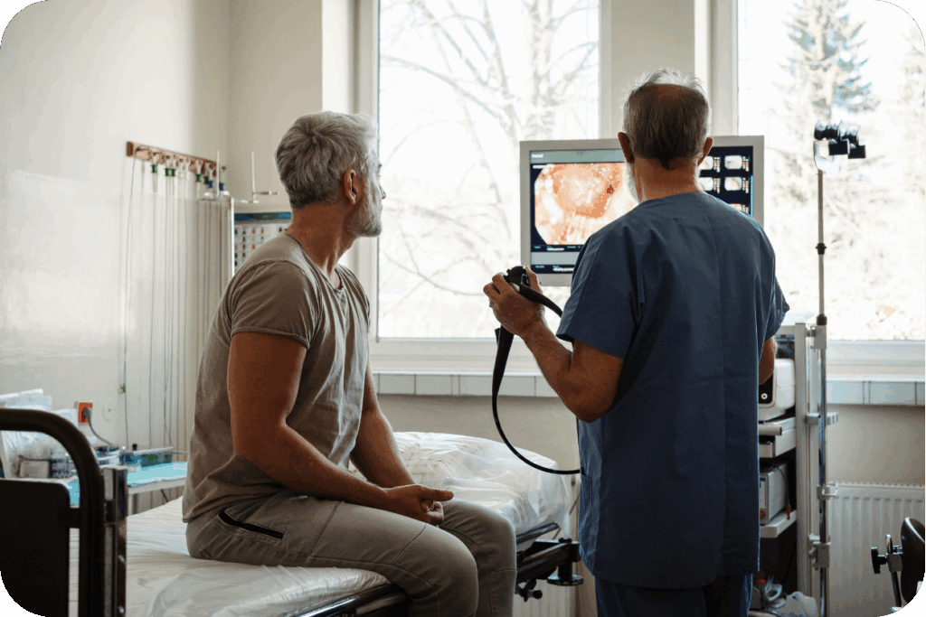 A imagem mostra um cenário clínico, onde um paciente (homem de cabelos grisalhos, sentado em uma maca) e um profissional de saúde (médico ou técnico, de costas e vestindo uniforme cirúrgico azul) observam juntos um monitor. O monitor exibe uma imagem interna detalhada do trato digestivo, típica de um exame de endoscopia ou colonoscopia, que o profissional está manipulando com um aparelho. A cena ilustra a detecção precoce de condições de saúde.